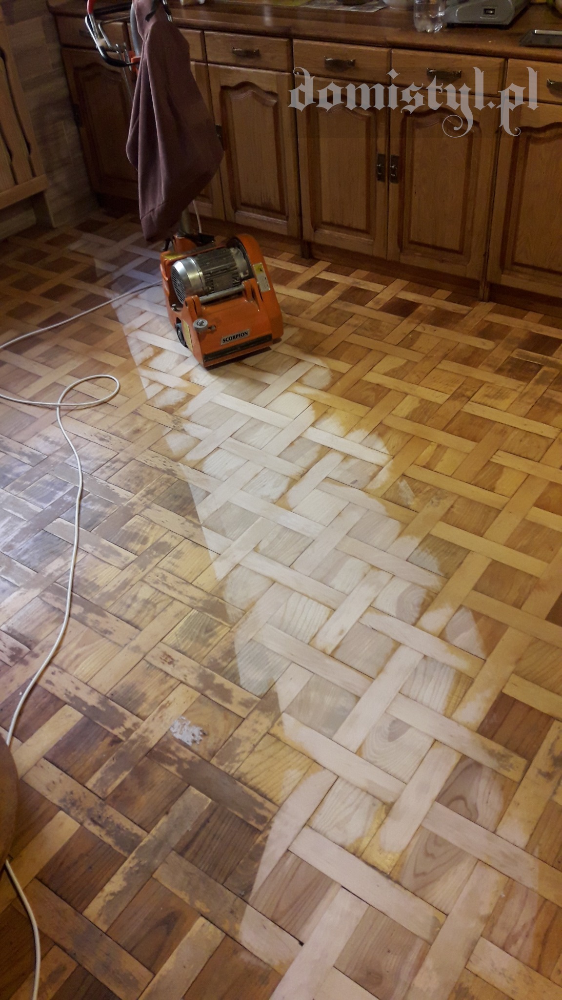 Parkiet w jodełkę częściowo odnowiony, widoczna różnica między starą a wyszlifowaną powierzchnią, obok stoi pomarańczowa szlifierka do parkietu marki Scorpion.