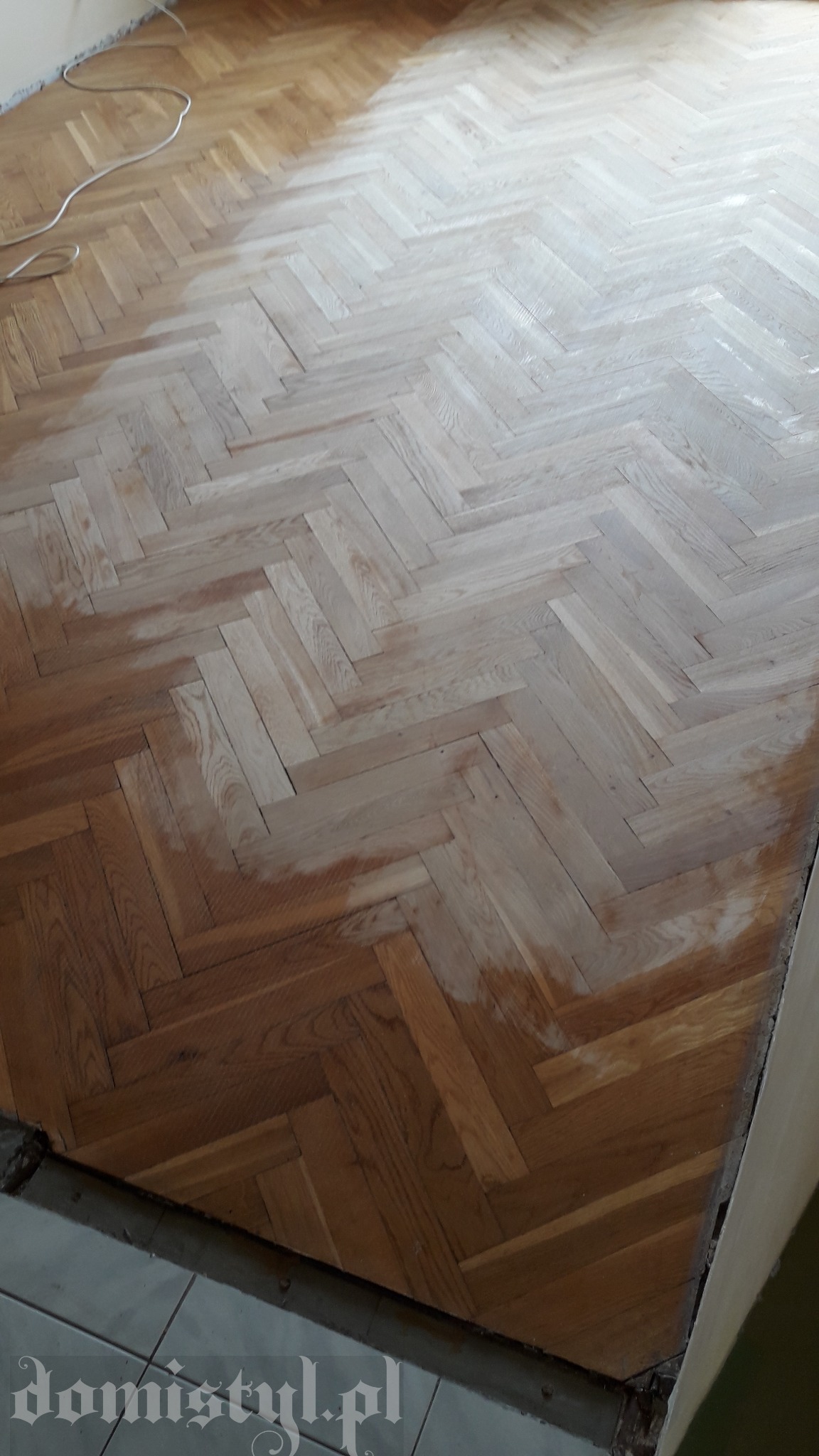 Parkiet ułożony w jodełkę, częściowo po renowacji, z widocznym kontrastem między odnowioną a starą powierzchnią, kabel przy ścianie.