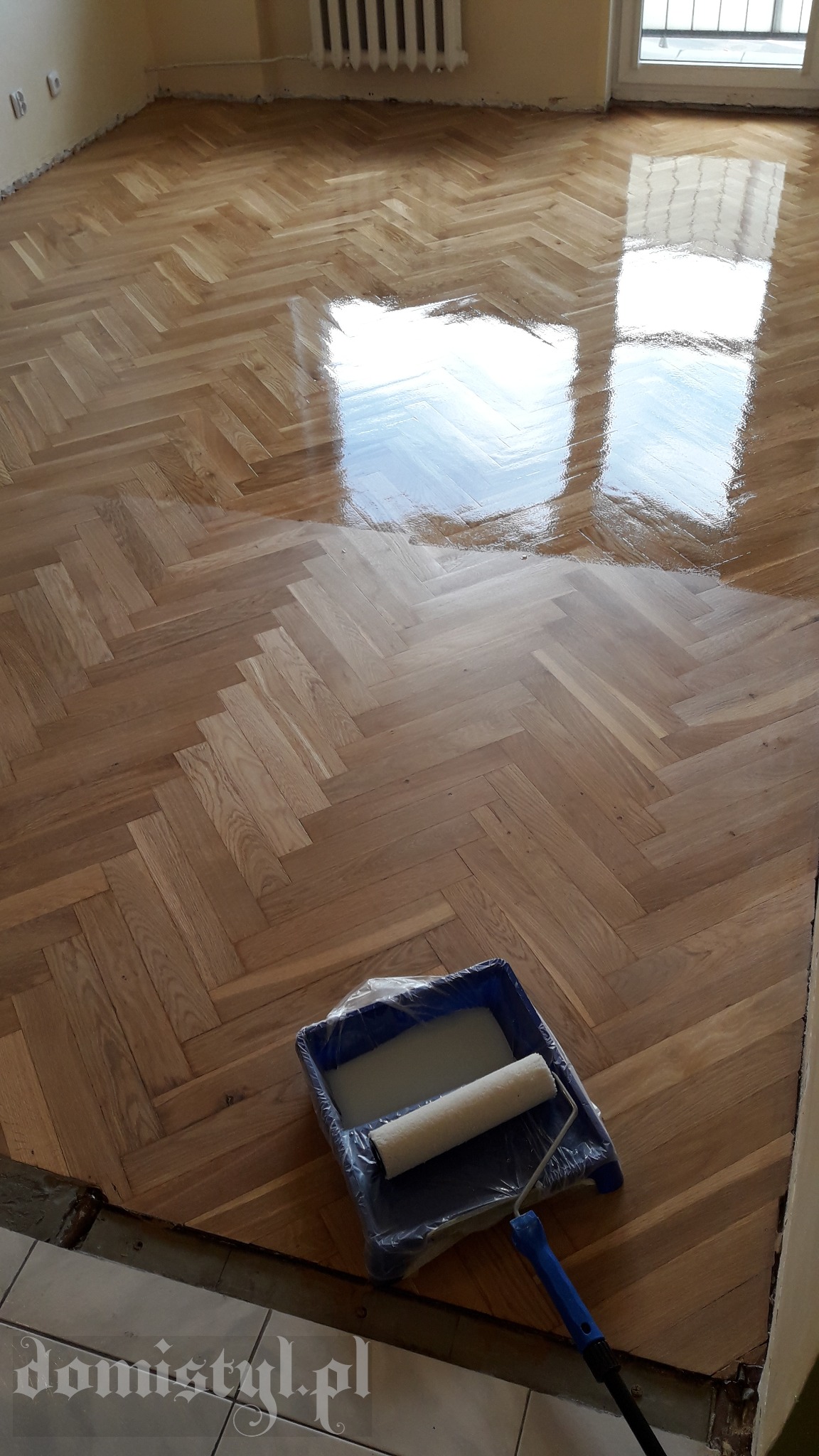 Świeżo polakierowany parkiet ułożony w jodełkę, z widocznym odbiciem okna, oraz kuweta z wałkiem do lakierowania w rogu pomieszczenia.
