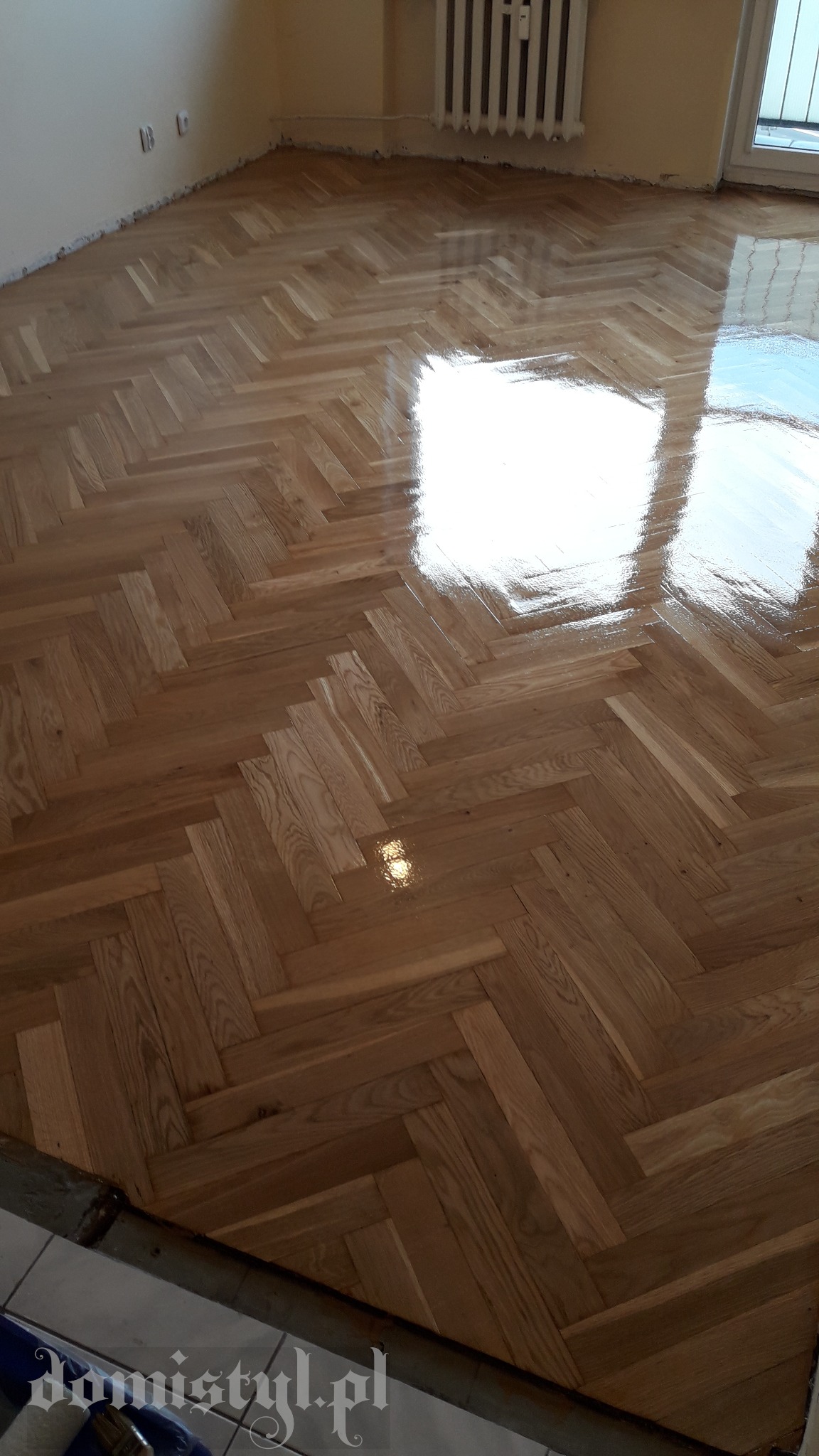Świeżo polakierowany parkiet ułożony w jodełkę, odbijający światło okna, widoczne ślady po taśmie malarskiej przy ścianie.