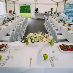 Uroczyste nakrycie stołu w białym namiocie: białe obrusy, kwiatowe dekoracje, zielone jabłka, elegancka zastawa i krzesła z białymi pokrowcami, widok na zielony trawnik.