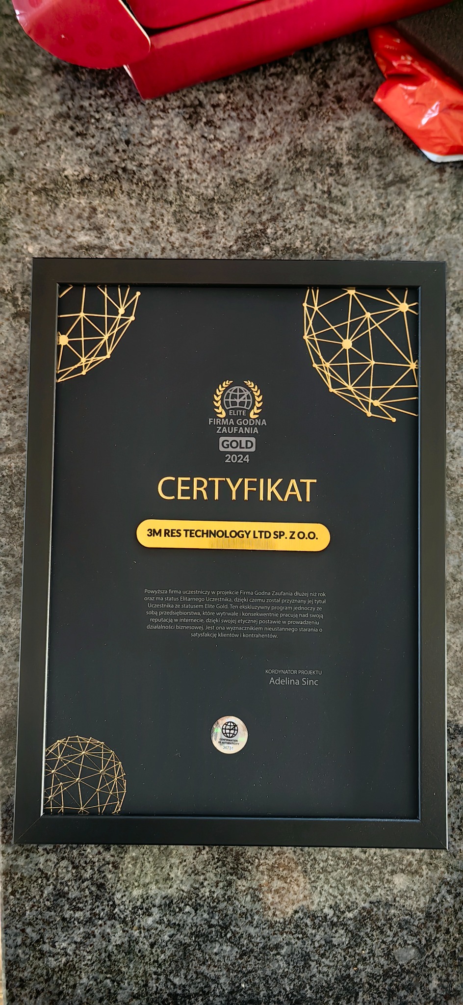 Oprawiony certyfikat 'Elite Firma Godna Zaufania Gold 2024' dla firmy 3M RES TECHNOLOGY LTD SP. Z O.O., leżący na szarym, marmurkowym tle.