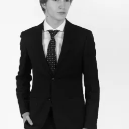 Czarno-białe zdjęcie młodego mężczyzny w czarnym garniturze i krawacie w kropki, pozującego na białym tle. Ujęcie od pasa w g&oacute;rę, z ręką w kieszeni.