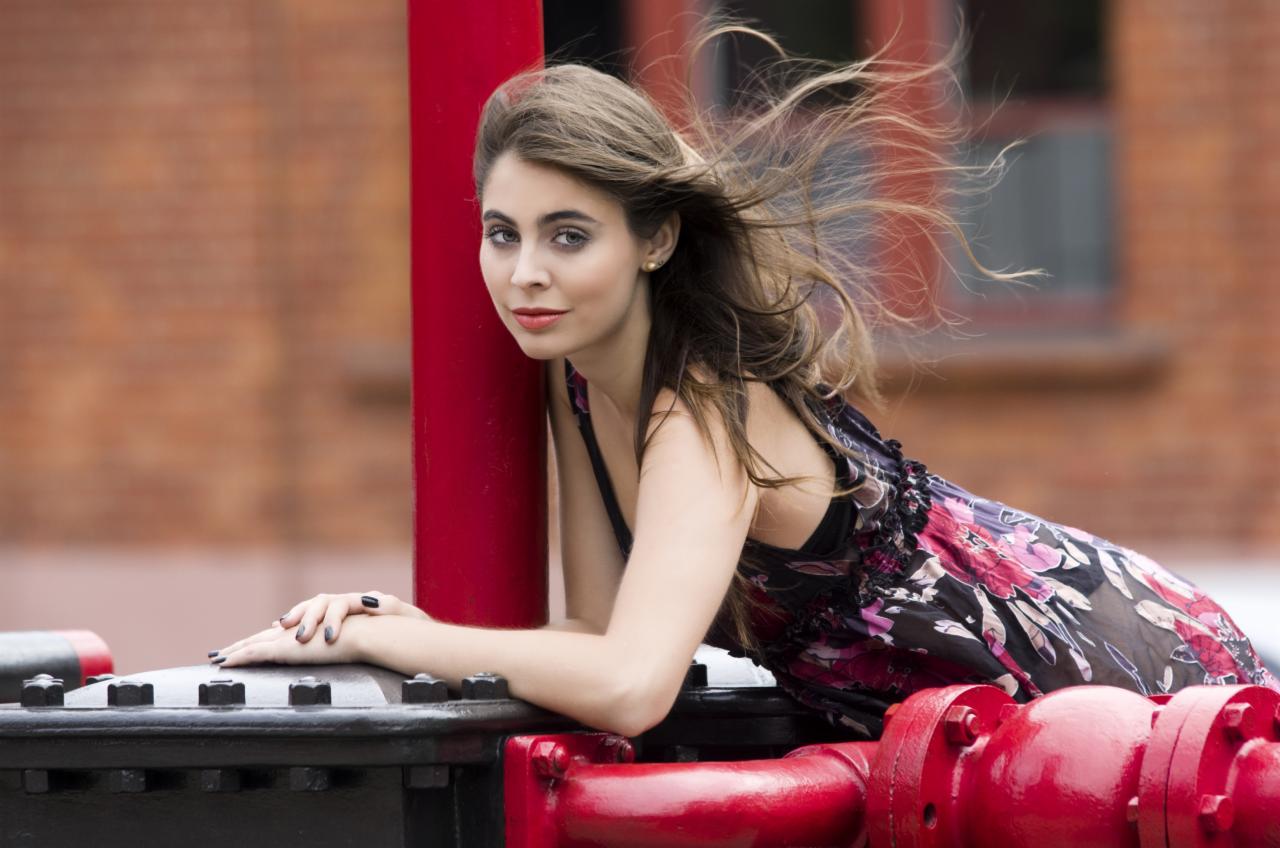 Portret kobiety w kwiecistej sukience, opierającej się o czerwoną konstrukcję na tle ceglanej ściany. Włosy rozwiane na wietrze, delikatny makijaż, naturalne światło.