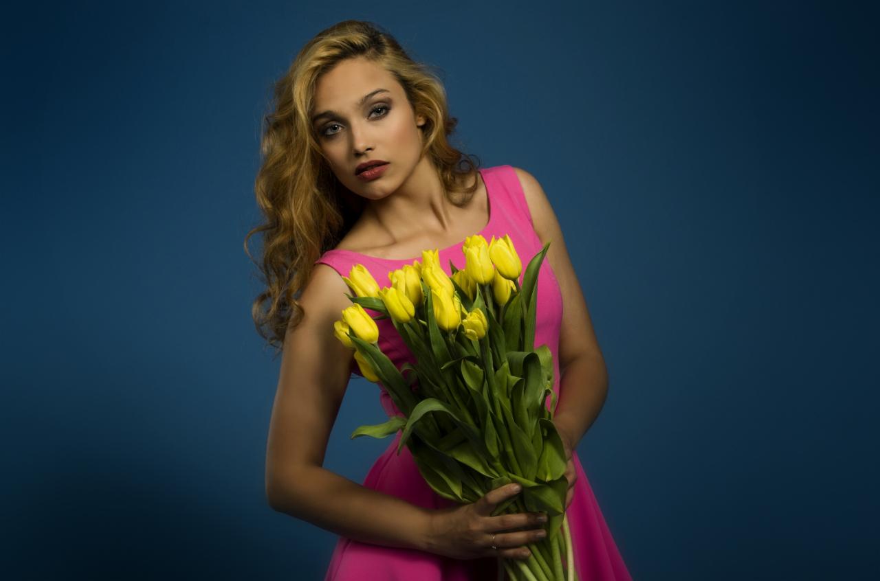 Portret kobiety w różowej sukience, trzymającej bukiet żółtych tulipanów na tle ciemnoniebieskiego studia. Subtelny makijaż i naturalne światło.