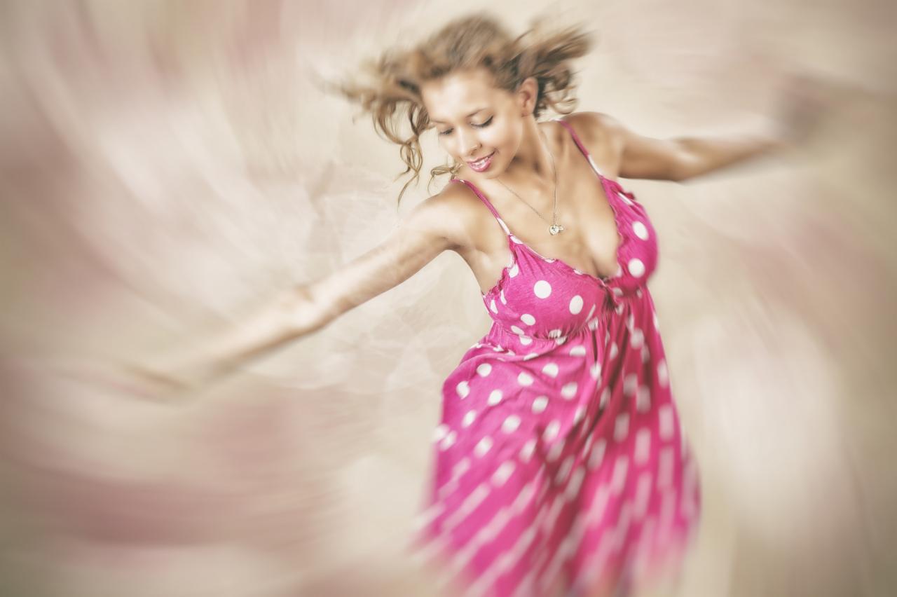 Rozmyte ujęcie kobiety w różowej sukience w białe grochy, z rozpostartymi ramionami, delikatny uśmiech, w ruchu, na rozmytym tle. Subtelna biżuteria na szyi.