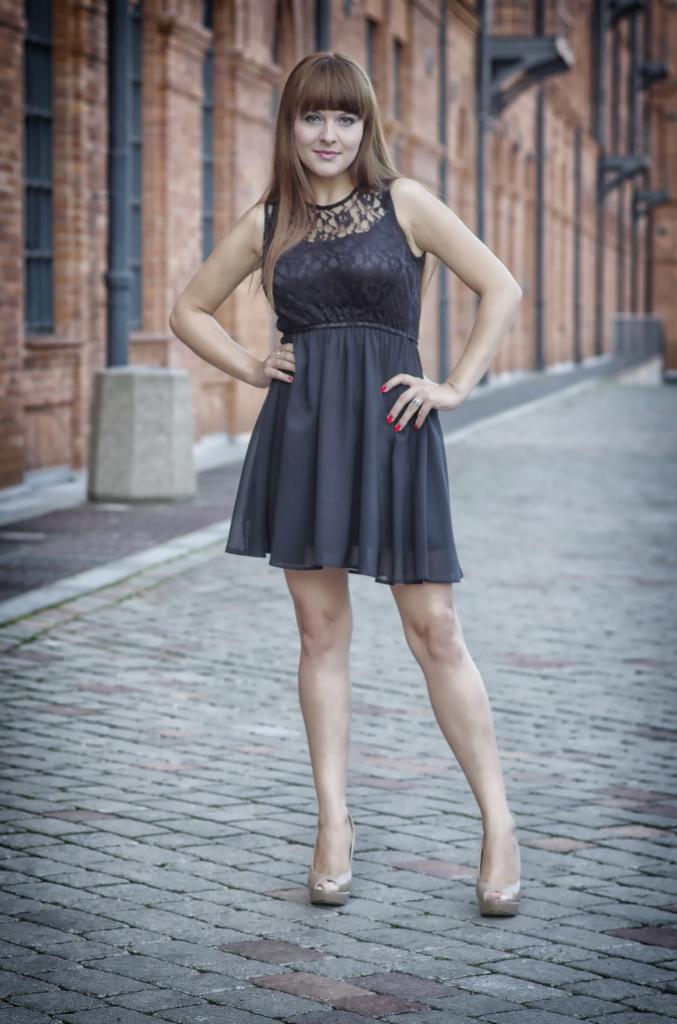Kobieta w czarnej sukience pozuje na brukowanej ulicy, z ceglanym budynkiem w tle. Uwagę zwracają czerwone paznokcie i beżowe szpilki.