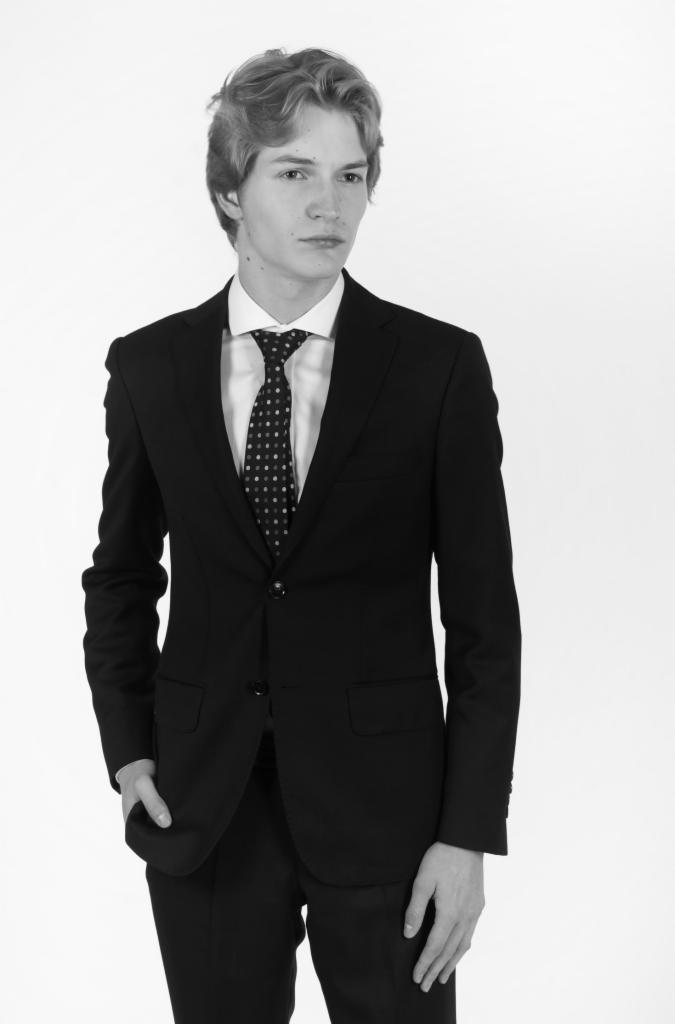 Czarno-białe zdjęcie młodego mężczyzny w czarnym garniturze i krawacie w kropki, pozującego na białym tle. Ujęcie od pasa w górę, z ręką w kieszeni.