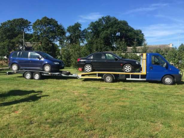 Niebieska laweta z dwoma samochodami: granatowym vanem i czarnym sedanem, zaparkowana na trawniku w słoneczny dzień.