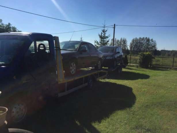 Dwa samochody osobowe zabezpieczone na platformie lawety, widoczne z boku, w tle zielona trawa i drzewa.