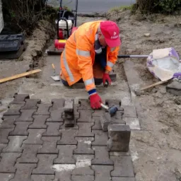 Pracownik w pomarańczowej kurtce i czerwonej czapce układa kostkę brukową na przygotowanym podłożu, widoczna taczka z piaskiem, ubijarka i narzędzia.