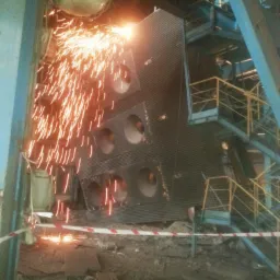 Demontaż konstrukcji stalowej palnikiem, iskrzenie. Fragment rusztowania i taśma ostrzegawcza w tle. Prace wyburzeniowe w hali przemysłowej, z bliska.