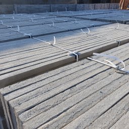 PROFIKS Ogrodzenia - Palety szarych, betonowych płyt prefabrykowanych, zabezpieczonych białymi taśmami, ułożone w stosy na placu budowy, z widocznym w tle fragmentem betoniarki.