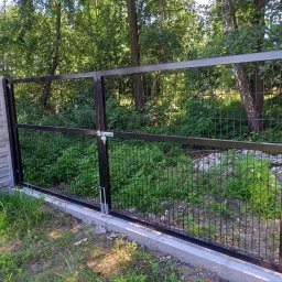 PROFIKS Ogrodzenia - Brama z ciemnych paneli ogrodzeniowych z zamkiem, zamontowana na betonowej podmurówce, przylegająca do ogrodzenia betonowego, w tle zieleń drzew.