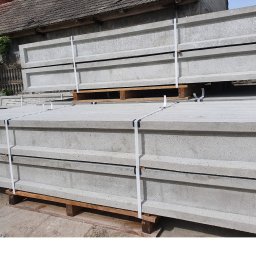 PROFIKS Ogrodzenia - Stos prefabrykowanych, betonowych elementów ogrodzeniowych na paletach, zabezpieczonych białymi taśmami, w tle budynek z szarą dachówką.