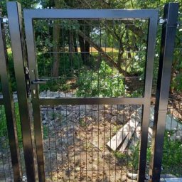 PROFIKS Ogrodzenia - Czarna furtka z paneli ogrodzeniowych z widocznym zamkiem i klamką, osadzona na betonowej podmurówce, w tle zieleń drzew.