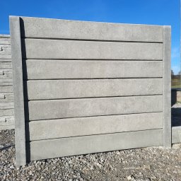 PROFIKS Ogrodzenia - Panel ogrodzeniowy betonowy z poziomymi wstawkami, widoczny na żwirowym podłożu, z boku ułożone kolejne panele, błękitne niebo w tle.
