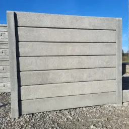 Panel ogrodzeniowy betonowy z poziomymi wstawkami, widoczny na żwirowym podłożu, z boku ułożone kolejne panele, błękitne niebo w tle.