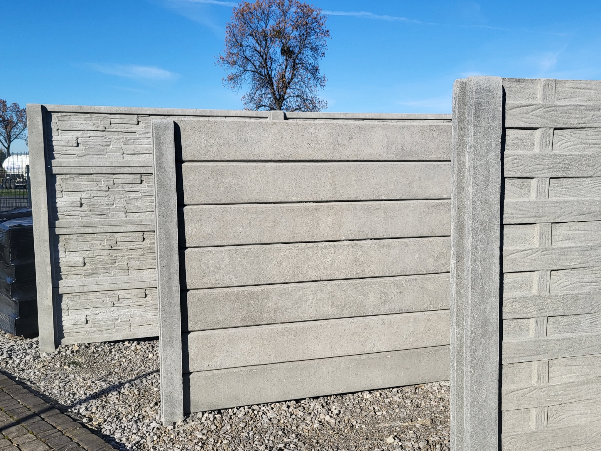 Fragment betonowego ogrodzenia z elementami dekoracyjnymi imitującymi kamień i drewno, widoczne słupki i panele na żwirowym podłożu, w tle drzewo bez liści pod błękitnym niebem.