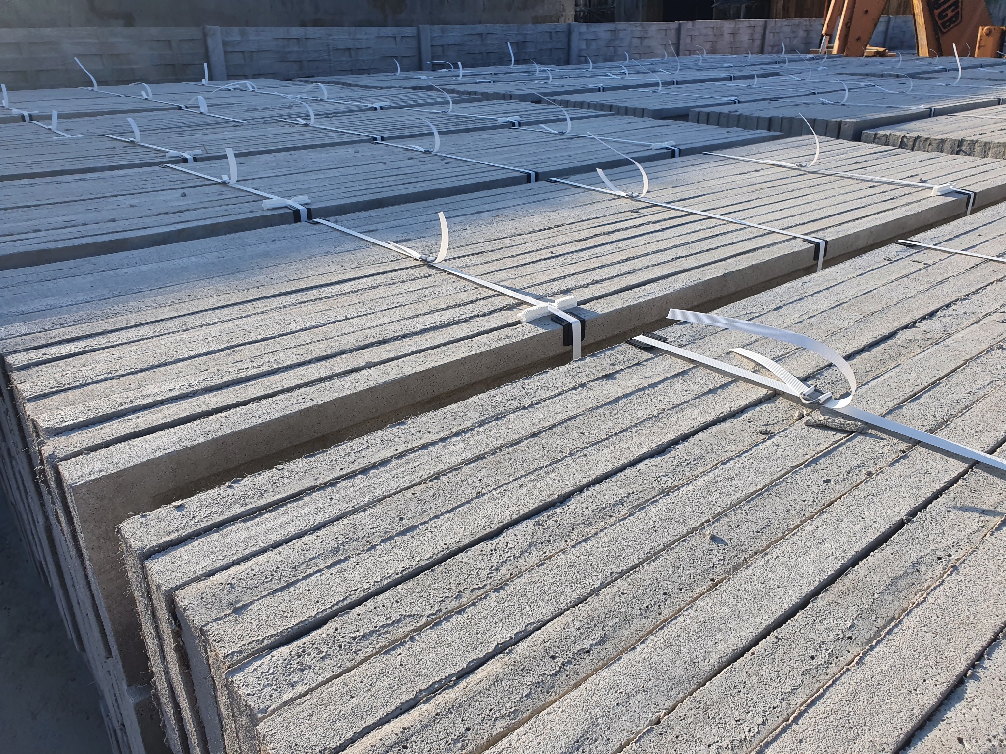 Palety szarych, betonowych płyt prefabrykowanych, zabezpieczonych białymi taśmami, ułożone w stosy na placu budowy, z widocznym w tle fragmentem betoniarki.