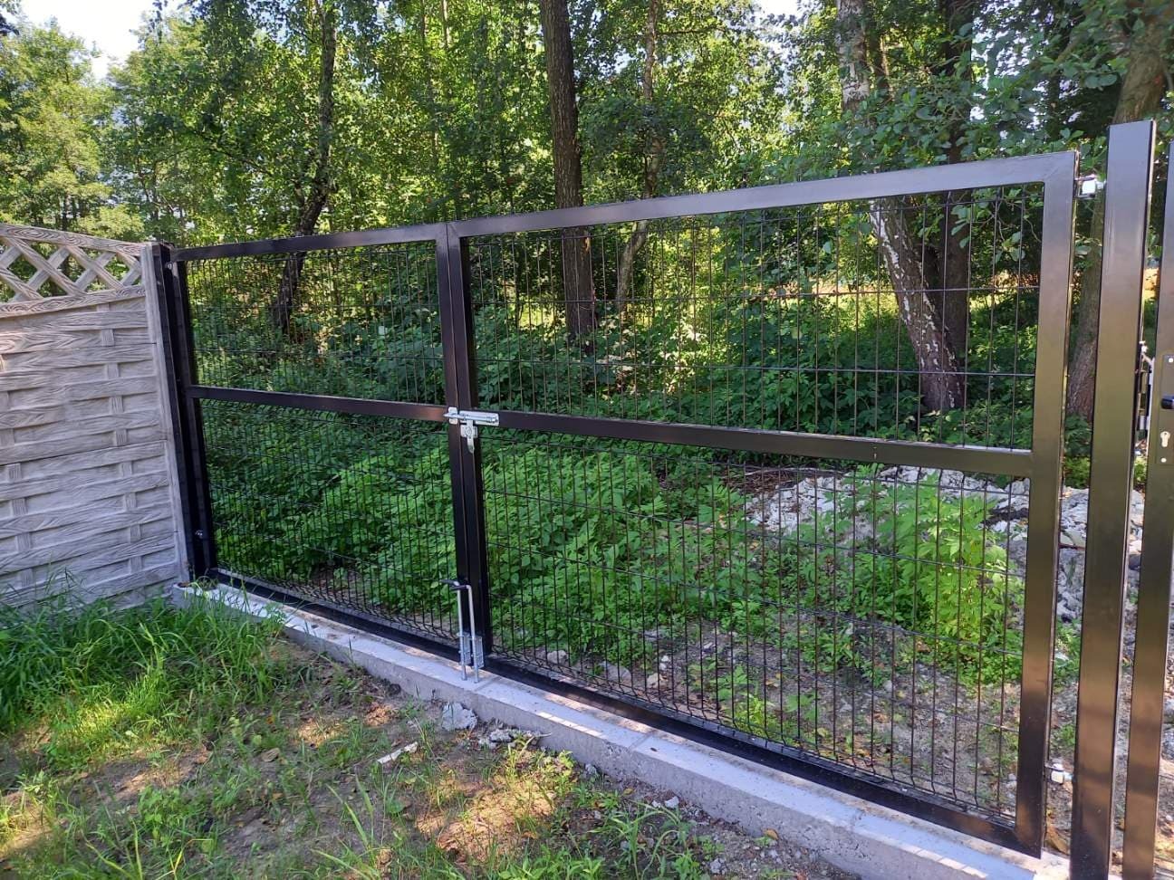 Brama z ciemnych paneli ogrodzeniowych z zamkiem, zamontowana na betonowej podmurówce, przylegająca do ogrodzenia betonowego, w tle zieleń drzew.
