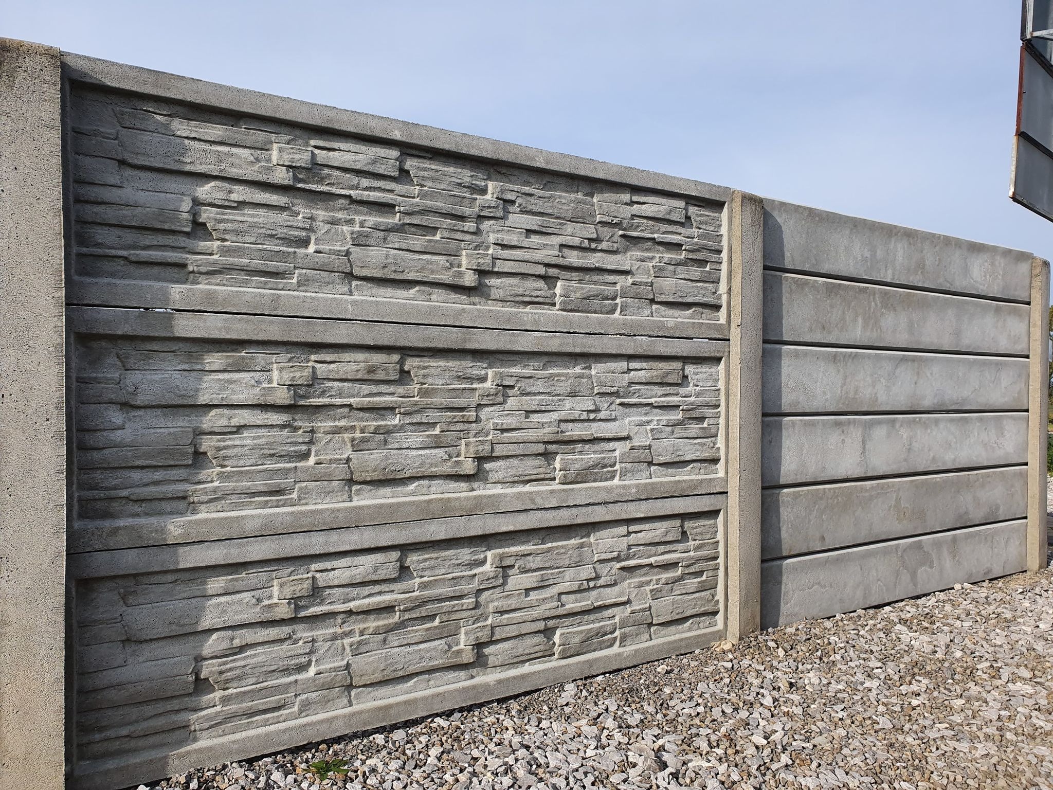 Szary, betonowy płot z prefabrykatów o fakturze imitującej kamień łupany, widoczny na tle nieba i żwirowego podłoża.
