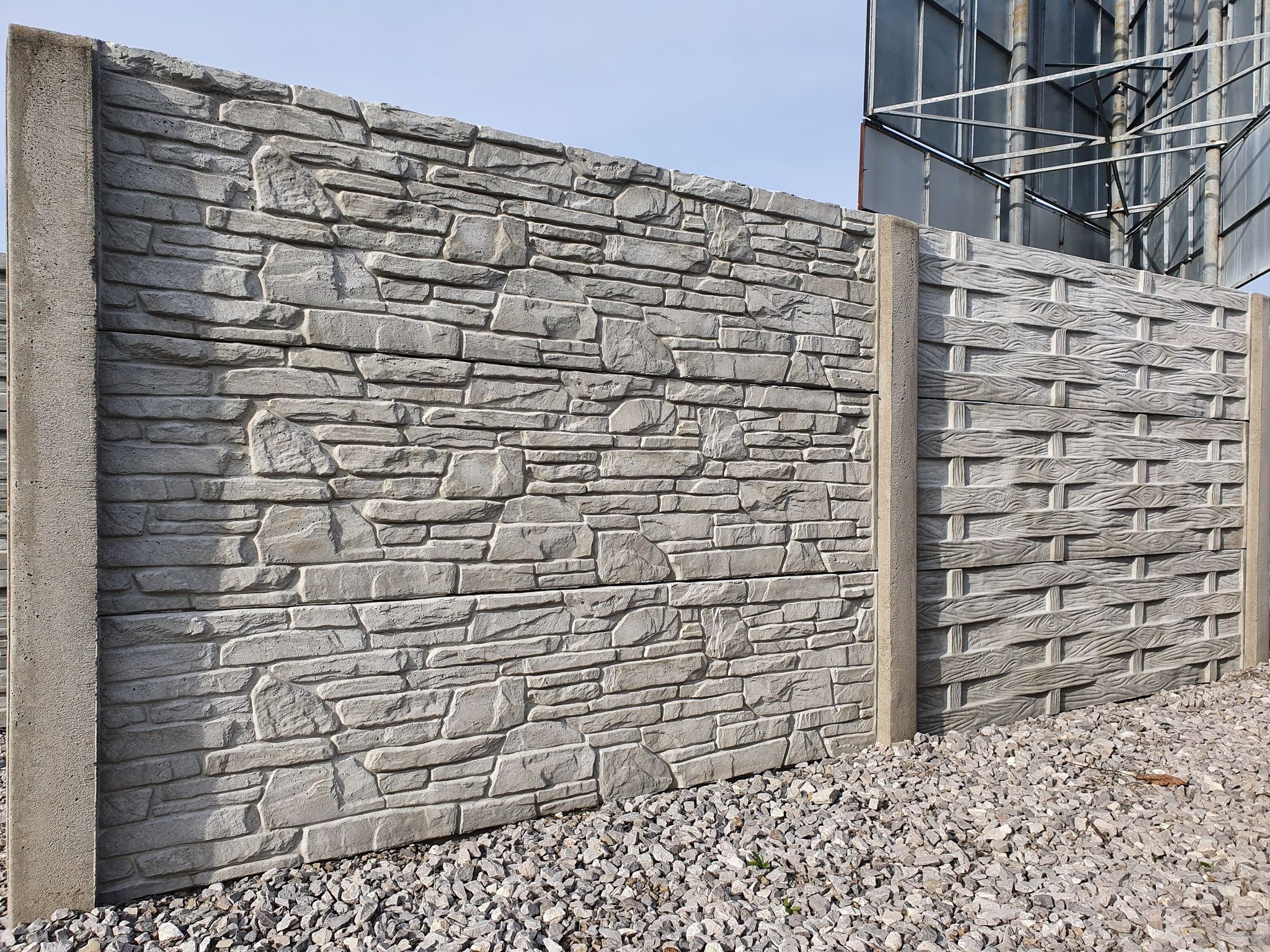 Dwa panele ogrodzeniowe z betonu: jeden imitujący kamień, drugi z wzorem plecionki, oddzielone słupem, na tle żwirowego podłoża i metalowej konstrukcji.