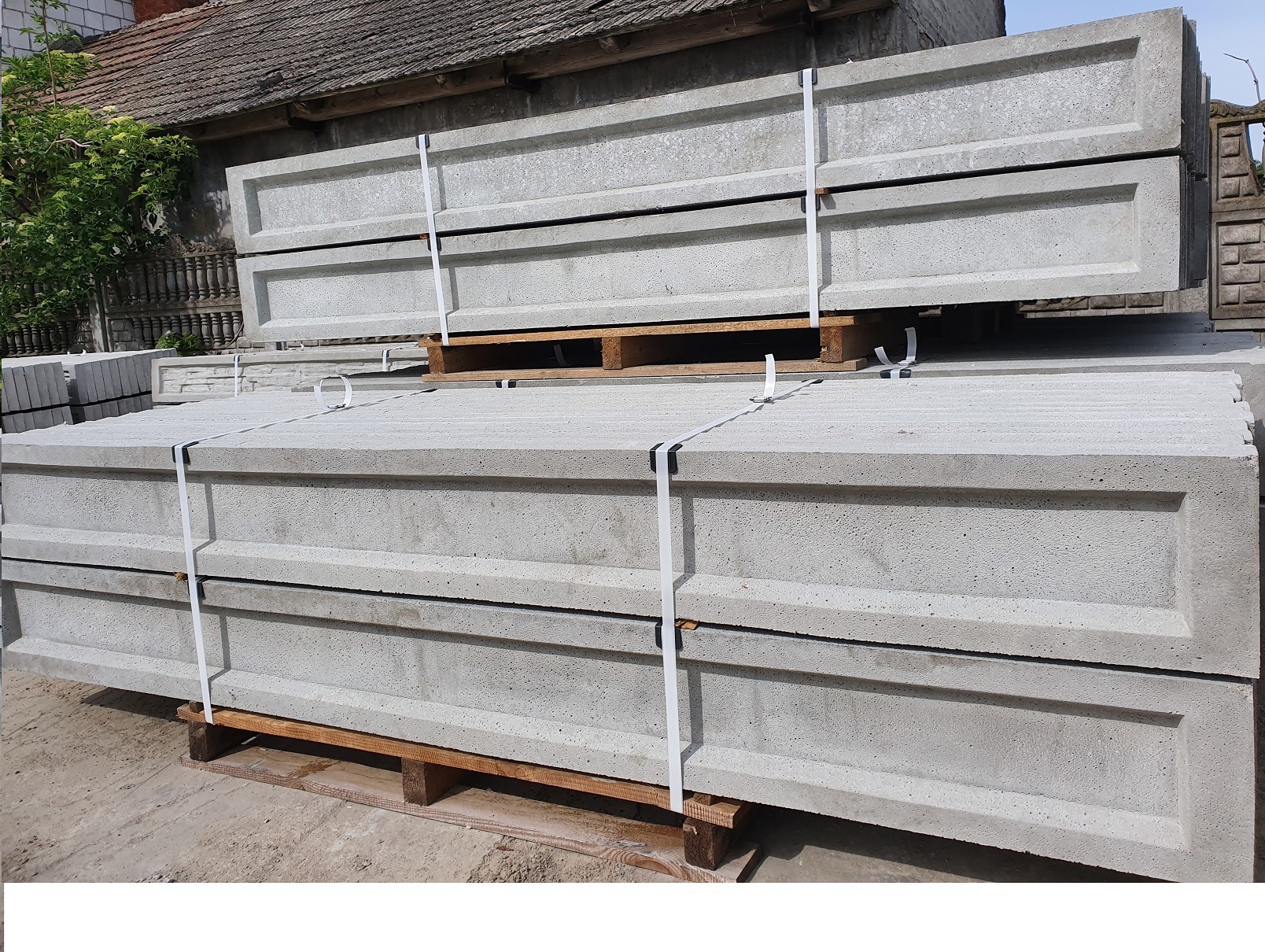Stos prefabrykowanych, betonowych elementów ogrodzeniowych na paletach, zabezpieczonych białymi taśmami, w tle budynek z szarą dachówką.