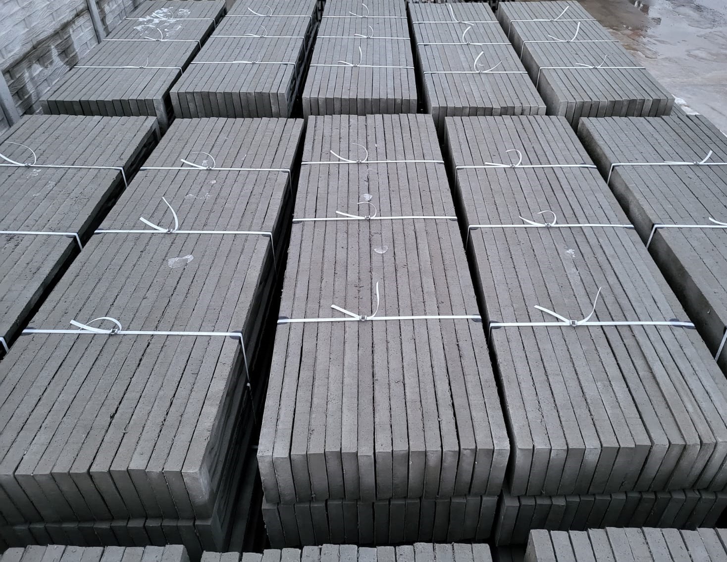 Szare płyty betonowe ułożone w stosy i zabezpieczone białymi taśmami, widoczne rowki na krawędziach płyt.