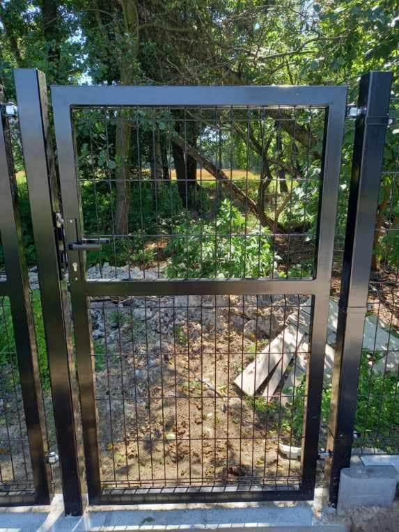 Czarna furtka z paneli ogrodzeniowych z widocznym zamkiem i klamką, osadzona na betonowej podmurówce, w tle zieleń drzew.