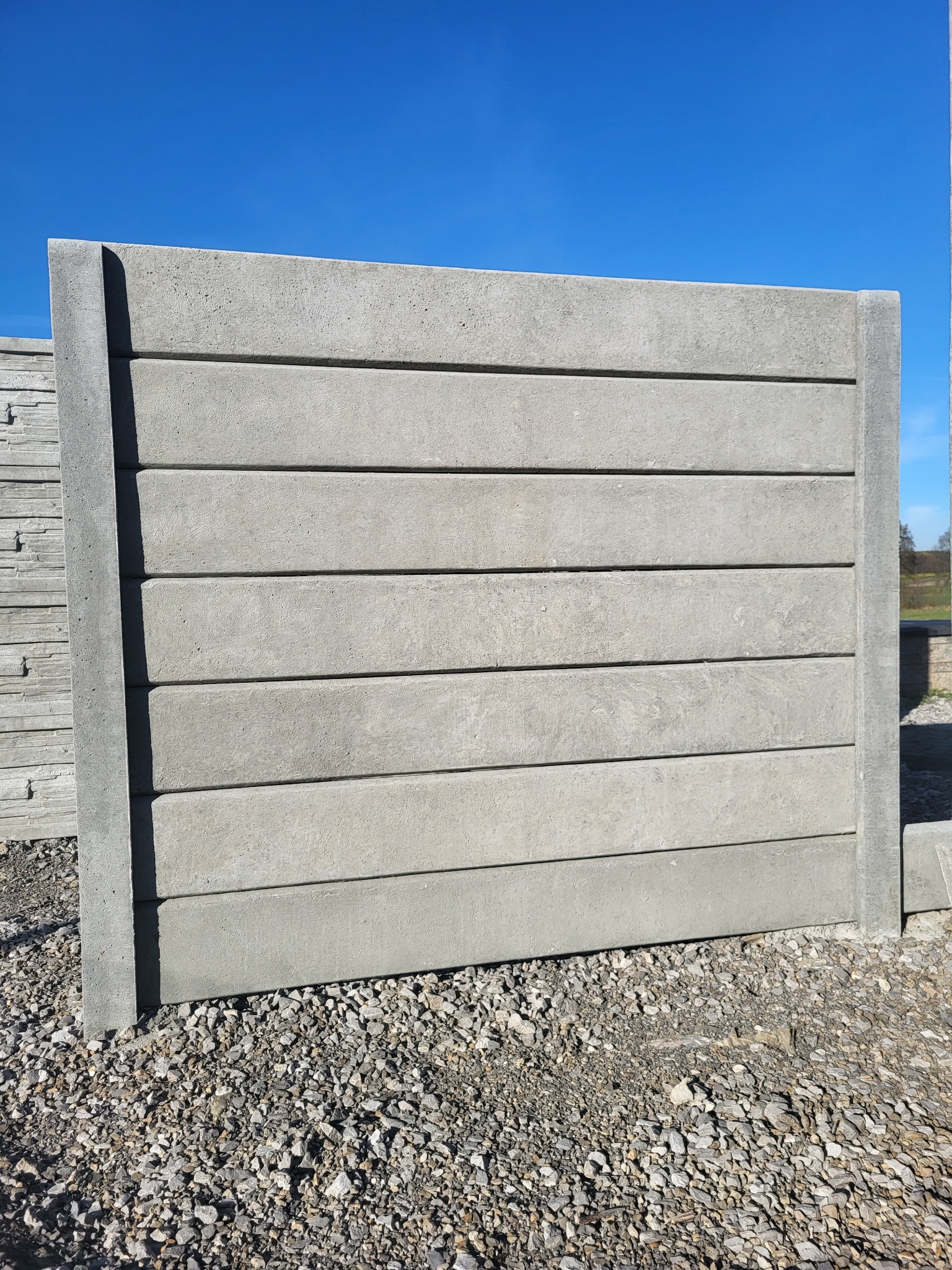 Panel ogrodzeniowy betonowy z poziomymi wstawkami, widoczny na żwirowym podłożu, z boku ułożone kolejne panele, błękitne niebo w tle.