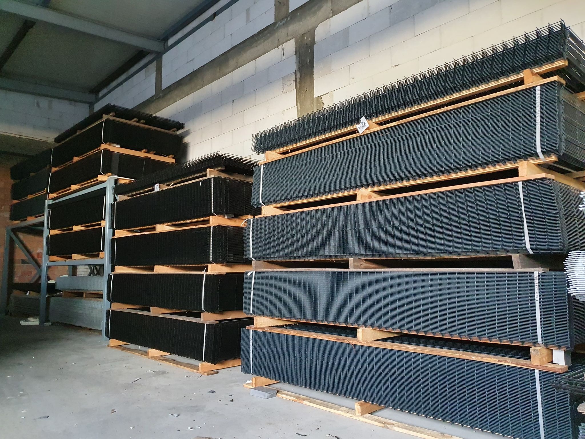 Magazyn z paletami wypełnionymi panelami ogrodzeniowymi zgrzewanymi, zabezpieczonymi białymi taśmami, na tle ściany z białych pustaków.