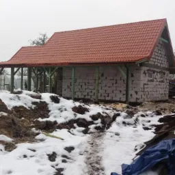 Wznoszony dom murowany z czerwonym dachem z dachówki, widoczne ślady śniegu i błota na placu budowy, w tle zaparkowany niebieski bus.