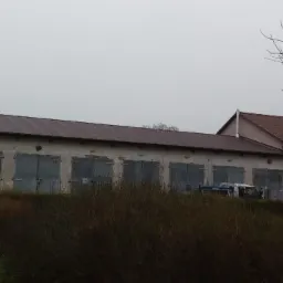 Długi budynek gospodarczy z brązowym dachem z blachy, widoczny zza krzewów, pochmurny dzień.