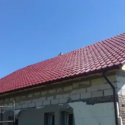 Czerwona dachówka ceramiczna na nowym domu w trakcie budowy, widoczne niezakończone mury i drewniana konstrukcja dachu, błękitne niebo w tle.