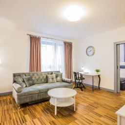 Oles Project Sp. Z o.o. - Jasne wnętrze mieszkania po remoncie: szara sofa z poduszkami, okrągły biały stolik kawowy, drewniana podłoga w jodełkę, biurko z krzesłem, widok na sypialnię z niebieskim łóżkiem.