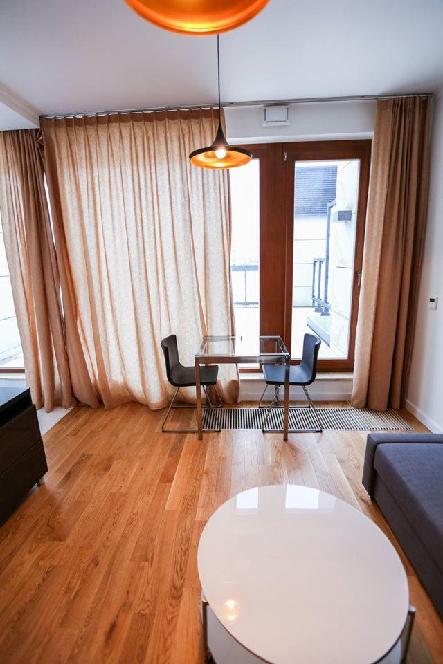 Jasne wnętrze pokoju z parkietem, beżowymi zasłonami i nowoczesnym oświetleniem, widok na balkon przez przeszklone drzwi.