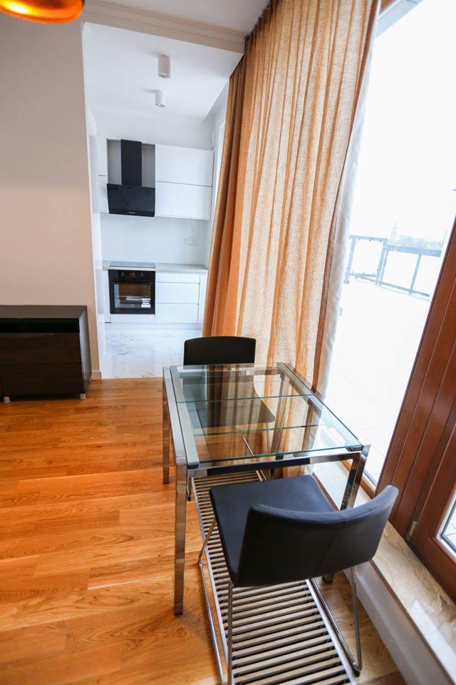 Jasne, nowocześnie urządzone mieszkanie z drewnianą podłogą, szklanym stołem i widokiem na kuchnię z białymi szafkami i czarnym okapem.