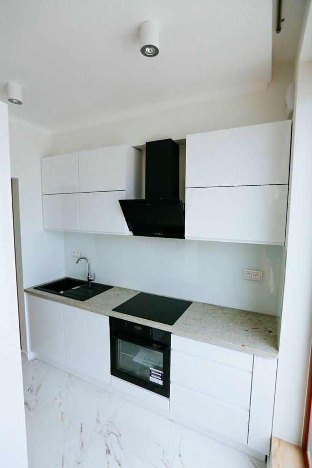 Nowoczesna, biała kuchnia z czarnym okapem i płytą indukcyjną, marmurowa podłoga i jasny blat, minimalistyczny design.