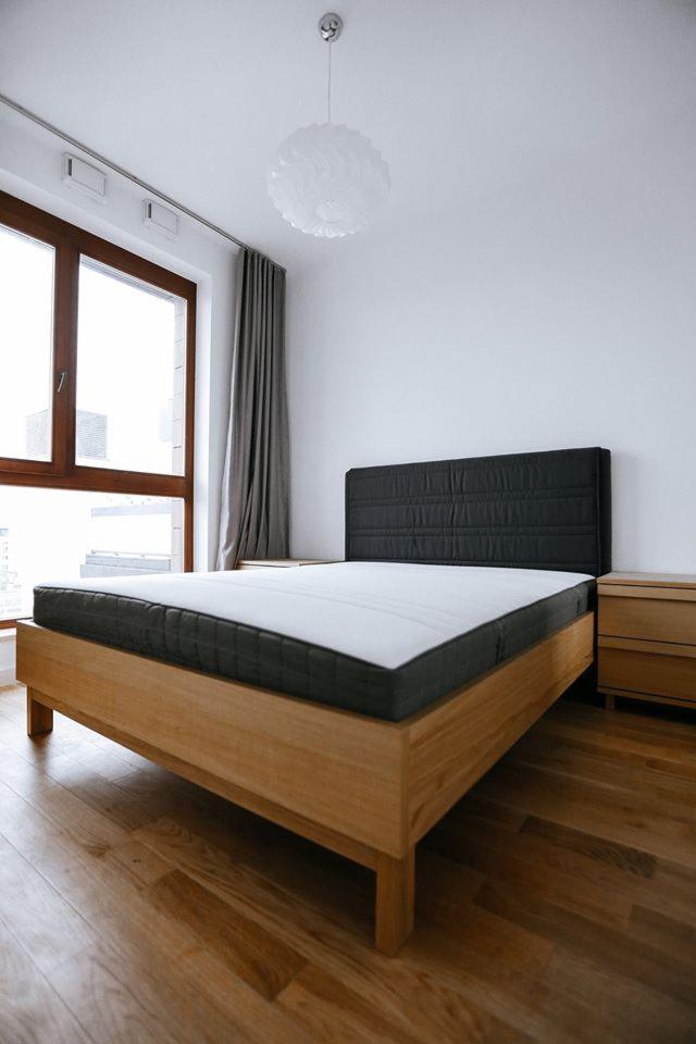 Jasne wnętrze sypialni z drewnianym łóżkiem z czarnym zagłówkiem i materacem, parkiet na podłodze, okno z szarymi zasłonami i lampa wisząca.