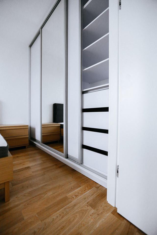 Nowoczesna szafa wnękowa z przesuwnymi, lustrzanymi drzwiami i otwartą częścią z półkami oraz szufladami z czarnymi uchwytami, na drewnianej podłodze w jasnym pomieszczeniu.