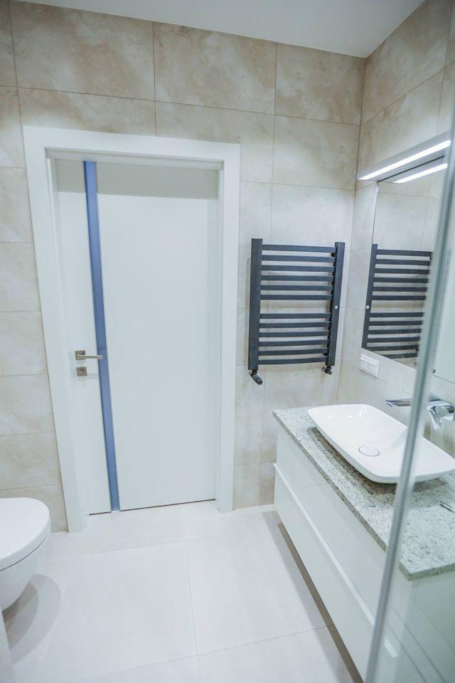 Nowoczesna łazienka z białymi płytkami, umywalką nablatową na szafce z szarym blatem, grzejnikiem drabinkowym w kolorze grafitowym oraz białymi drzwiami z niebieskim akcentem.