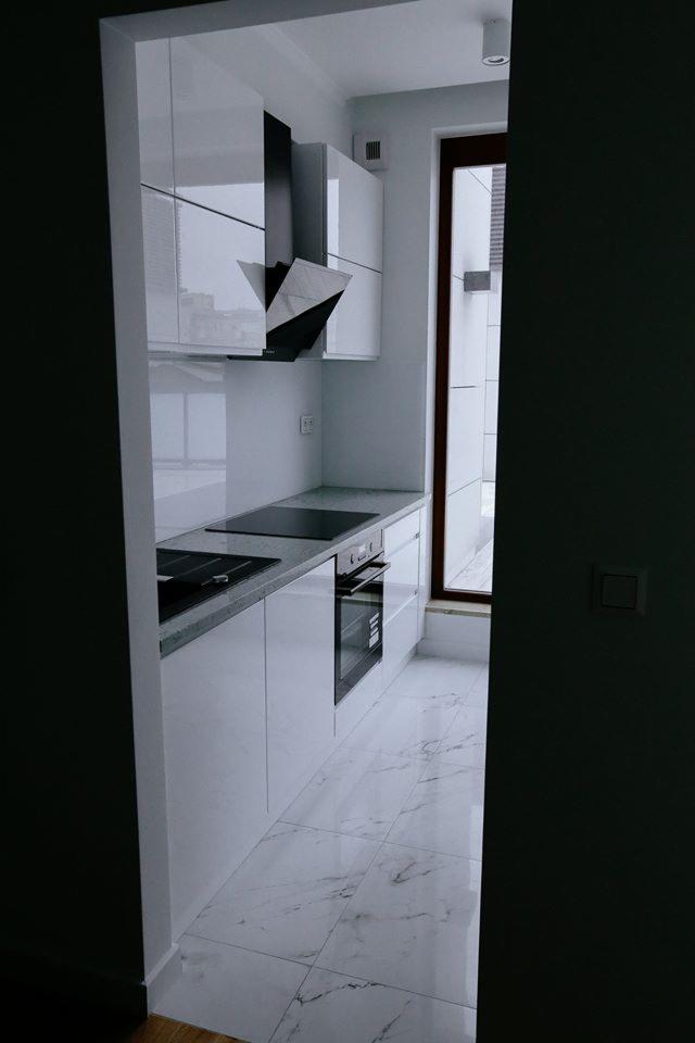 Nowoczesna, biała kuchnia z marmurową podłogą, czarnym okapem i indukcyjną płytą grzewczą, widok z korytarza, minimalistyczny design.