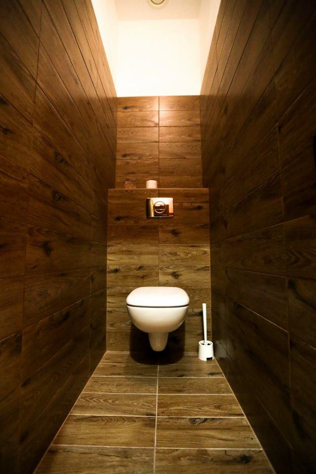 Wąska toaleta wykończona brązowymi płytkami imitującymi drewno, z podwieszaną miską WC i minimalistycznym przyciskiem spłukującym.