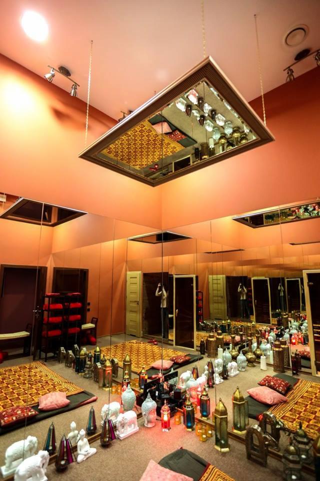 Pomieszczenie z pomarańczowymi ścianami, wyposażone w lustra, orientalne dywany, poduszki i dekoracyjne lampiony, z lustrzanym sufitem podwieszanym na łańcuchach.