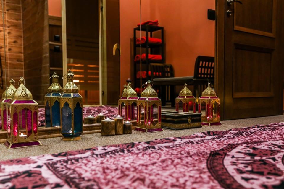 Dekoracyjne lampiony ze świecami w kolorach fioletu, złota i błękitu, ustawione na wzorzystym dywanie w pomieszczeniu z drewnianą sauną i półkami z ręcznikami w tle.