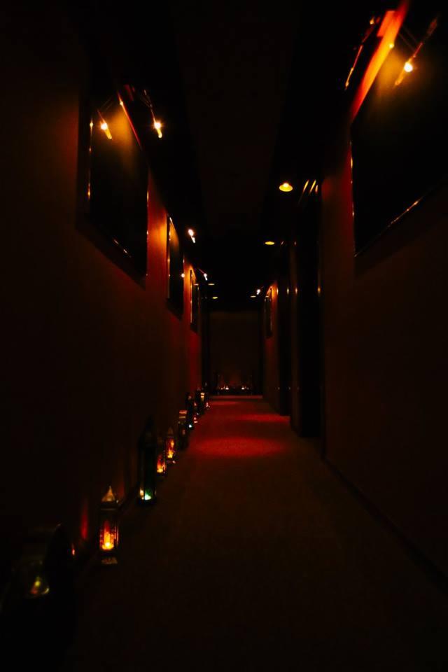Długi, wąski korytarz oświetlony nastrojowymi lampkami i lampionami, tworzącymi ciepłą, czerwoną poświatę na podłodze i ścianach. Ściany w odcieniach ciemnej czerwieni, z ciemnymi ramami okiennymi...