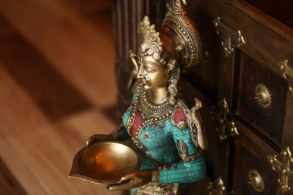 Dekoracyjna, pozłacana statuetka kobiety w turkusowo-czerwonym stroju, trzymająca miseczkę, ustawiona na tle drewnianej komody z metalowymi zdobieniami.