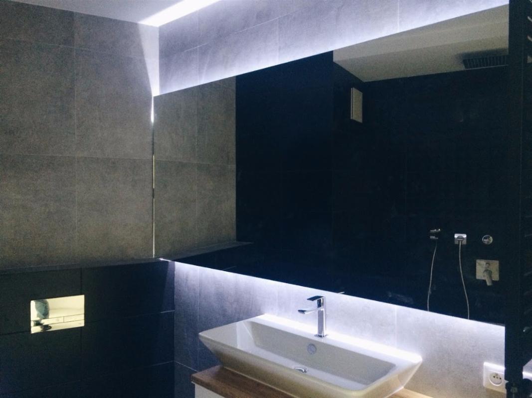 Nowoczesna łazienka z umywalką nablatową, podświetlanym lustrem i szarą glazurą, widoczne elementy instalacji wodnej i elektrycznej.