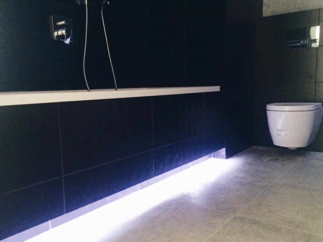 Nowoczesna łazienka z czarnymi płytkami, podświetleniem LED u dołu ściany i wiszącą toaletą.